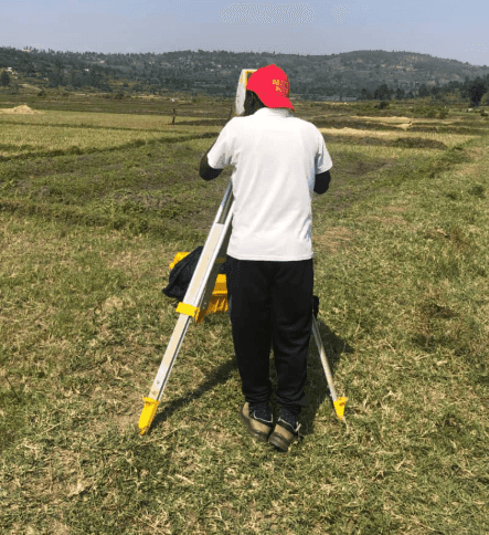 Slide 2 — A surveyor in a red cap stands behind a leveling instrument on a tripod in a lush green field, with hills in the background.