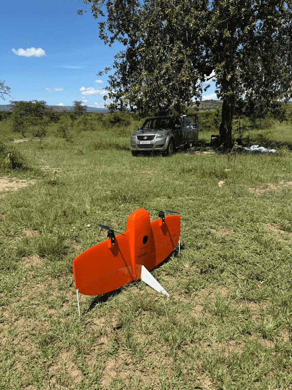 Slide 5 — An orange drone rests on grassy terrain beside a parked silver SUV under a tree, with blue skies and distant hills in the background.