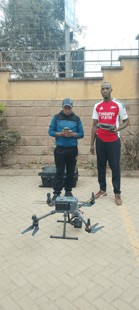 Slide 6 — Two individuals stand beside a drone on a paved surface, one checking a device while the other holds a controller, with a wall in the background.