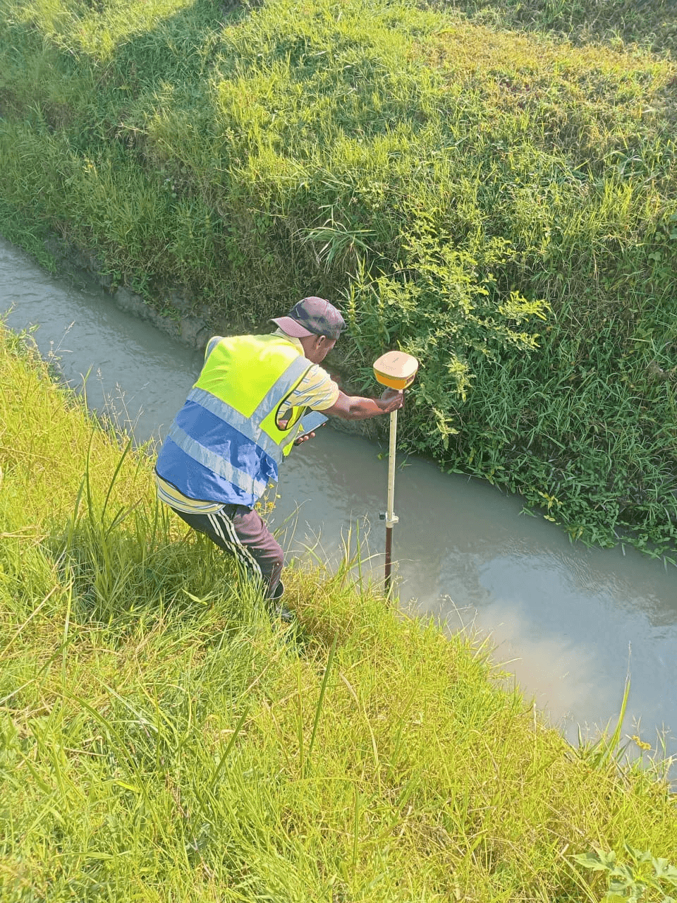 Slide 7 — A worker in a high-visibility vest uses surveying equipment near the edge of a grassy drainage canal.